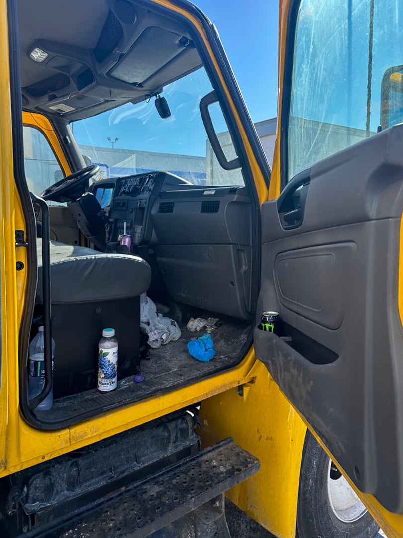 The interior of a yellow truck cab with the drivers door open, showing a cluttered floor with trash, bottles, and cans. Sunlight shines through the window, and part of the trucks exterior and wheel are visible.