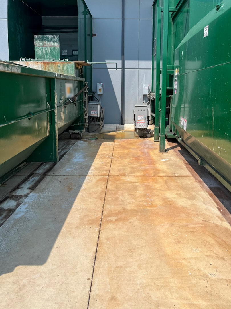 Two large green industrial dumpsters or compactors are placed on a concrete pad beside a building. Electrical boxes and wires are visible on the wall, and the ground shows rusty stains and marks.