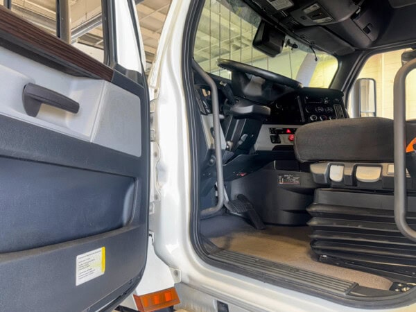 View of a trucks driver-side interior, showing the open door, steps, seat, dashboard, pedals, and steering wheel under bright indoor lighting.