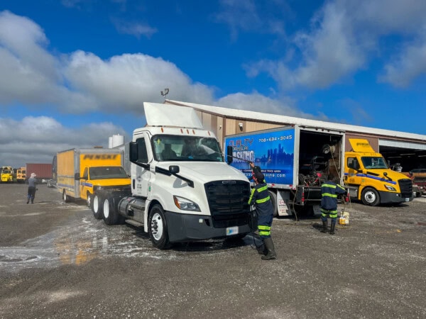 Workers in reflective uniforms wash a white semi-truck parked outside a large garage. Nearby are other yellow and white trucks, a service van, cleaning equipment, and blue sky with clouds overhead.