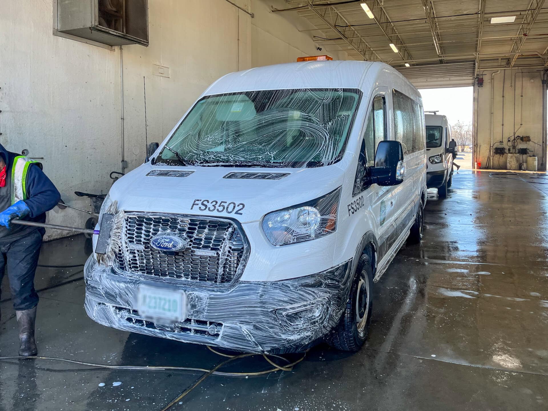A white Ford van is covered in clear plastic film, parked inside a large, wet, industrial garage. A worker in a reflective vest stands on the left side, partially out of frame. Another similar van is parked behind it.