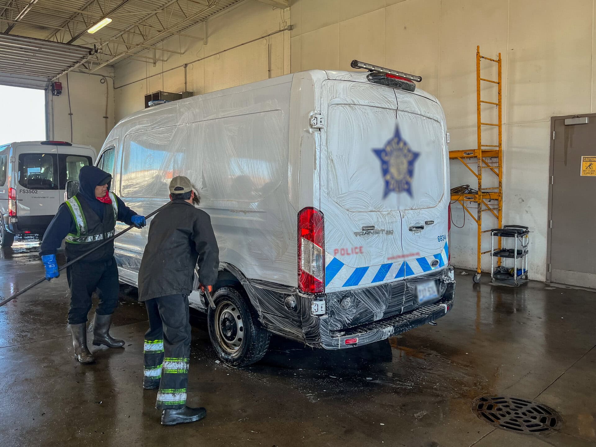 Two workers in safety gear wash a white police van inside a garage. The van is covered in soap suds, and cleaning supplies are on the floor. A yellow ladder leans against the wall in the background.