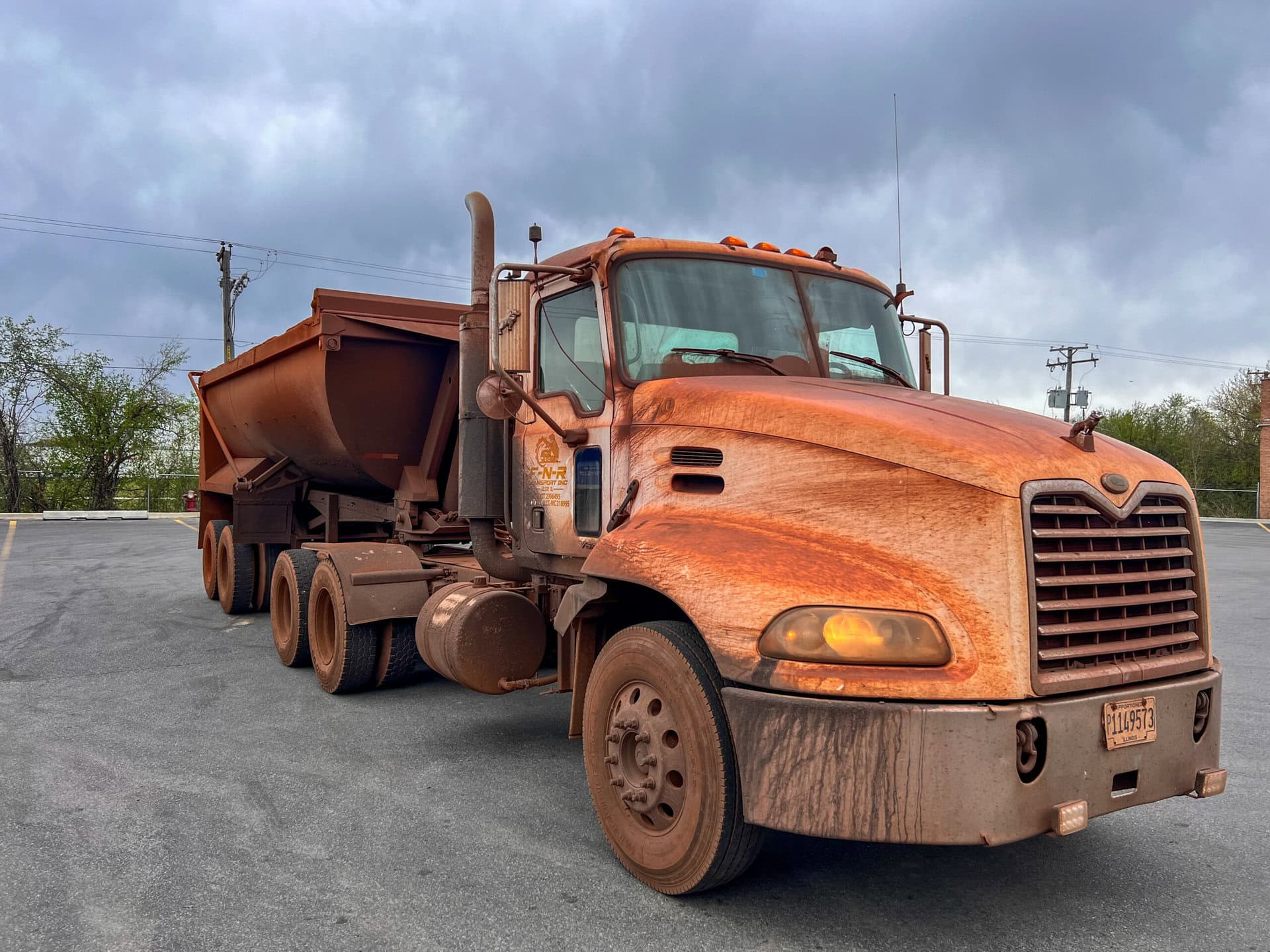 A large, rust-covered dump truck is parked on an empty lot under a cloudy sky, with visible dirt and mud on its exterior and wheels.