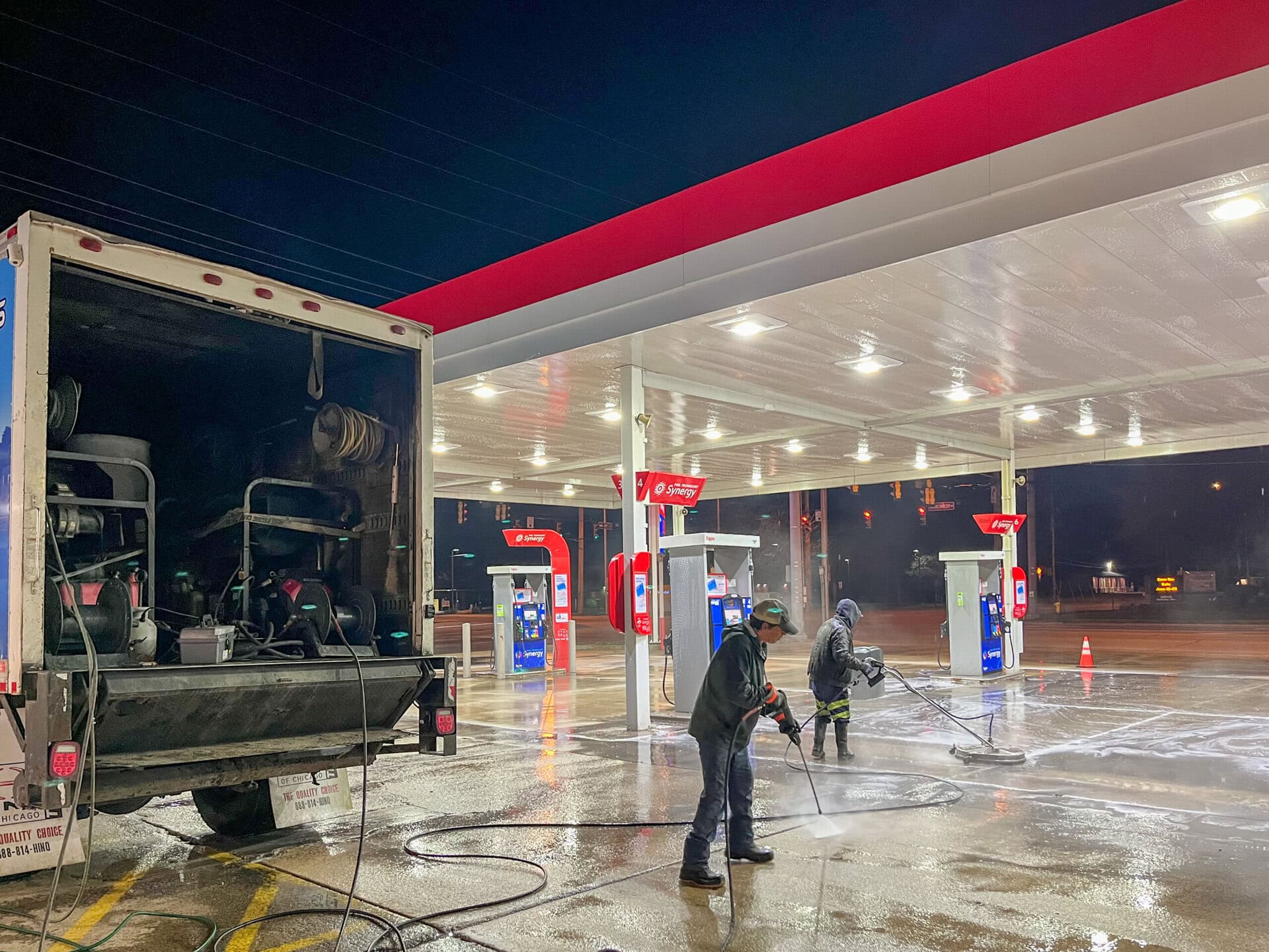 Two people use power washers to clean the ground at a gas station at night. A large truck with equipment is parked nearby, and the gas pumps are empty. The area is wet and reflects the lights above.