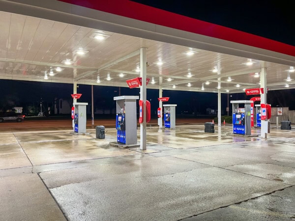A brightly lit gas station at night with several blue and red fuel pumps under a white canopy, standing on a wet concrete surface. No cars or people are visible.
