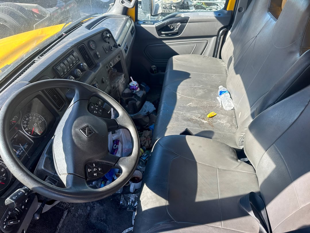 The interior of a worn, cluttered truck cab with black seats, a steering wheel, trash on the floor, and a plastic water bottle on the passenger seat. Sunlight streams in through the windows.
