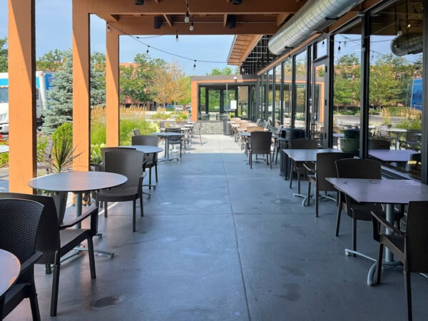 Outdoor restaurant patio with round tables and black chairs arranged along a covered walkway next to large windows. String lights hang above, and greenery and trees are visible in the background.