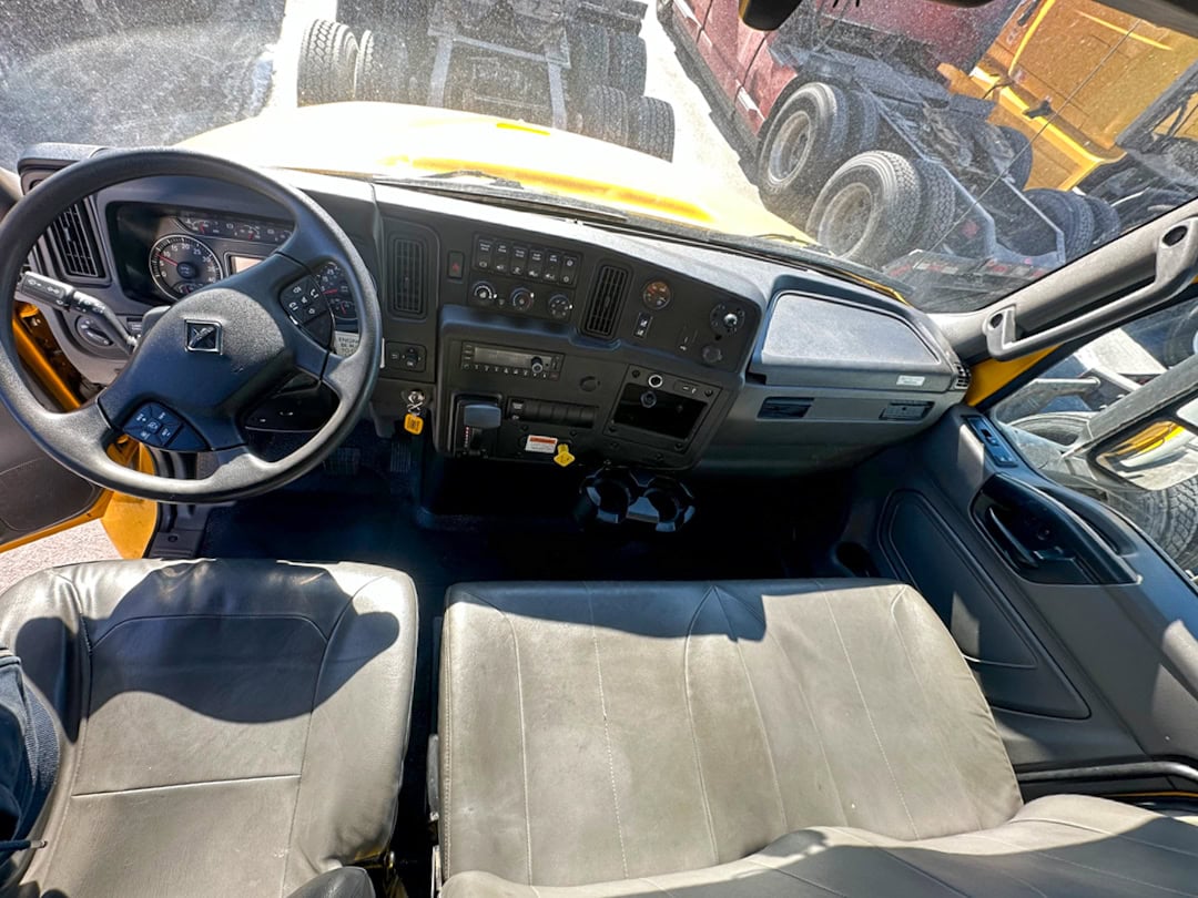 Wide-angle view of a school bus driver’s seat and dashboard, showing steering wheel, control panels, two front seats, and the windshield overlooking other parked buses outside.