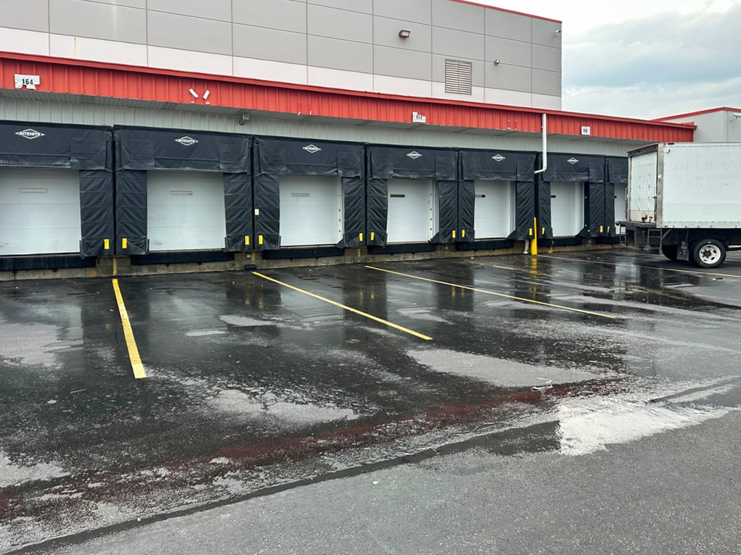 A row of five truck loading docks with black bumpers and closed bay doors next to a wet, reflective parking area. A white truck trailer is backed up to the dock on the far right.