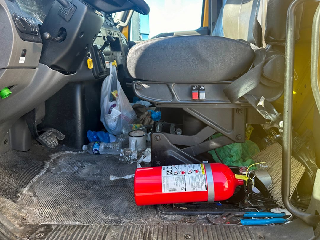 The interior of a vehicle cab with a cluttered floor, including a red fire extinguisher, plastic bottles, trash, and various items scattered beneath and around the drivers seat.