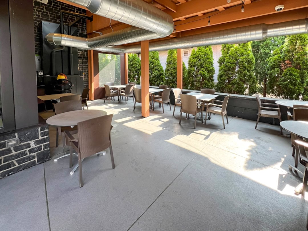 Outdoor patio seating area with round tables and brown chairs, under an orange wooden roof with exposed silver air ducts. Tall green shrubs line the background, and sunlight streams across the concrete floor.