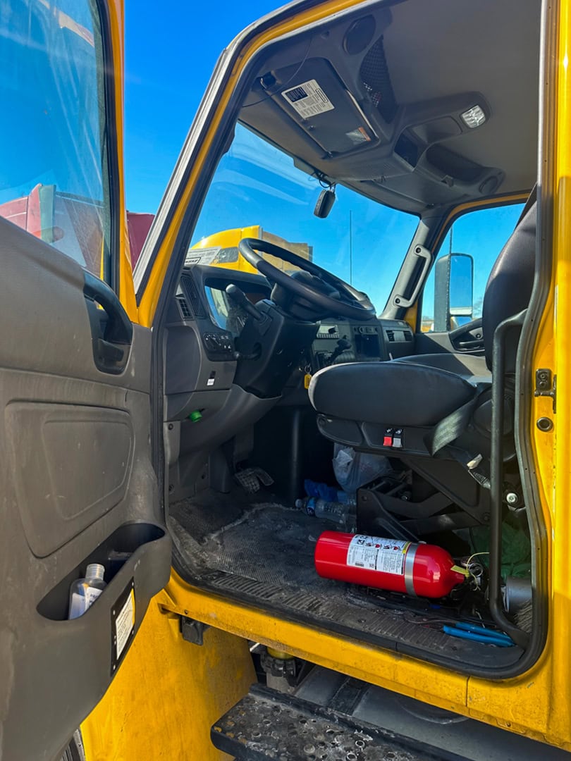 View of the interior of a yellow truck cab with the driver’s door open. A red fire extinguisher lies on the dusty floor near the driver’s seat. The dashboard, steering wheel, and controls are visible.