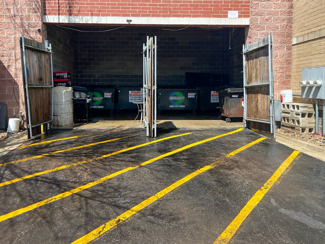 A gated outdoor garbage area with several dumpsters inside, surrounded by brick walls. The ground has bright yellow diagonal lines painted on the blacktop leading up to the open gates.