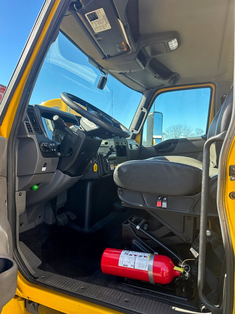 The interior of a yellow truck cab, showing the drivers seat, steering wheel, dashboard, and a red fire extinguisher secured on the floor near the seat.