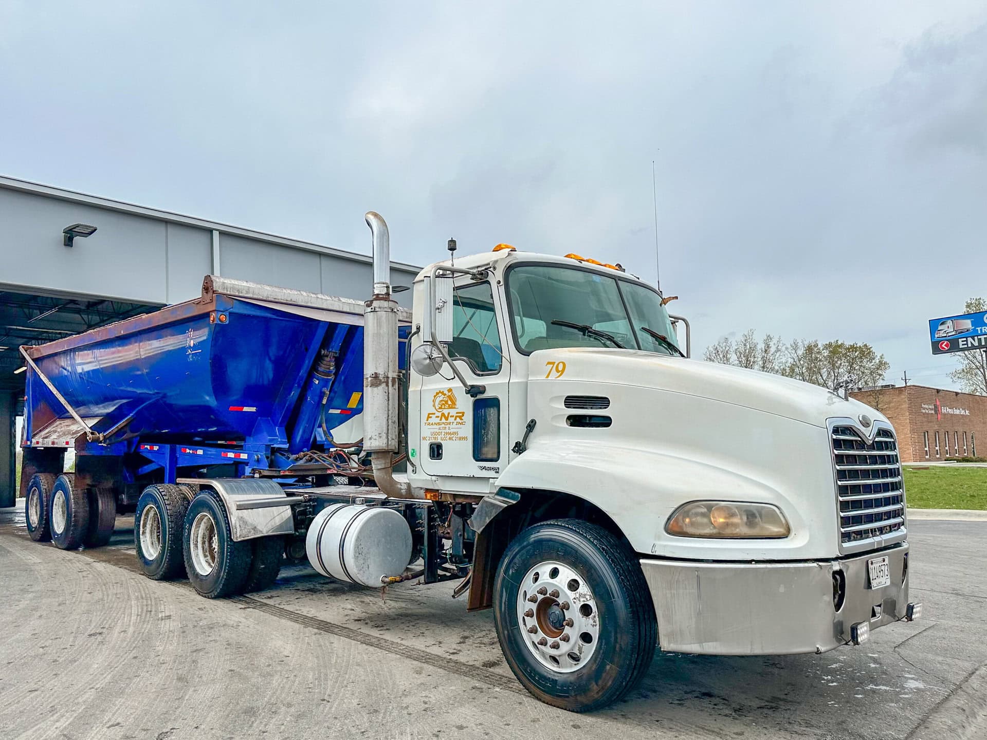 A white semi-truck with a blue dump trailer is parked outside near a building and a brick structure under a cloudy sky. The truck has the number 79 and a company logo on the door.