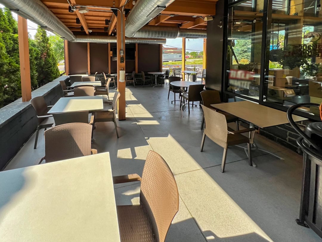A covered outdoor patio at a restaurant with several empty tables and chairs arranged neatly. Sunlight streams in from one side, and greenery is visible along the edge. The area appears clean and inviting.