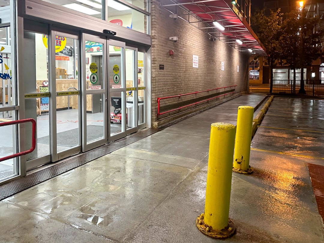 A well-lit store entrance at night with automatic glass doors, yellow bollards, red railings, and a wet concrete sidewalk reflecting the lights.