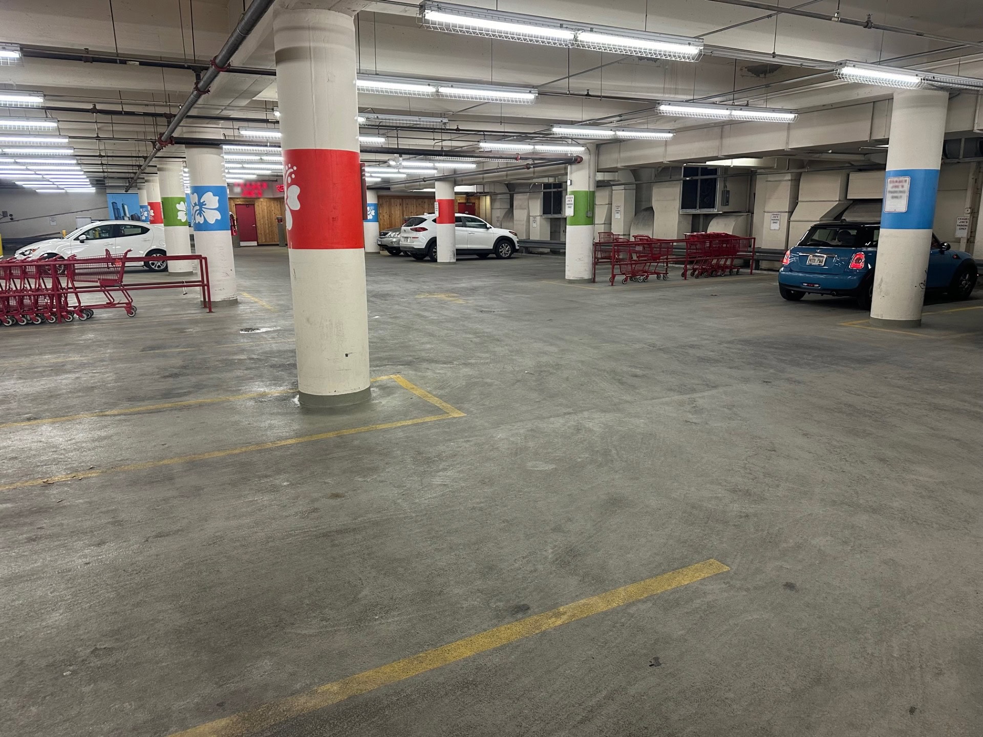 An indoor parking garage with concrete floors and pillars, several parked cars, red shopping cart returns, and colorful pillar signs. The lighting is fluorescent, and the garage appears mostly empty.