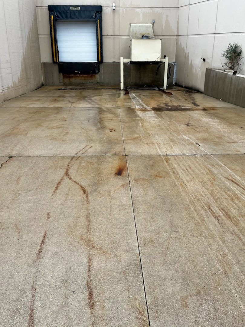 A concrete loading dock area with rust stains on the ground, a closed white overhead door with a black and yellow dock shelter, and a large metal container against a beige building wall.