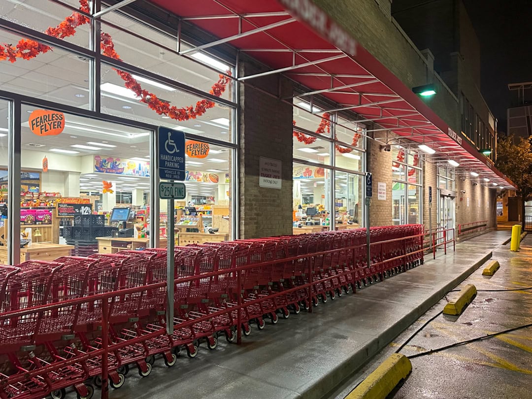 A row of red shopping carts lines the sidewalk outside a brightly lit grocery store at night, with glass windows displaying the interior and orange decor above. A handicapped parking sign is visible near the entrance.