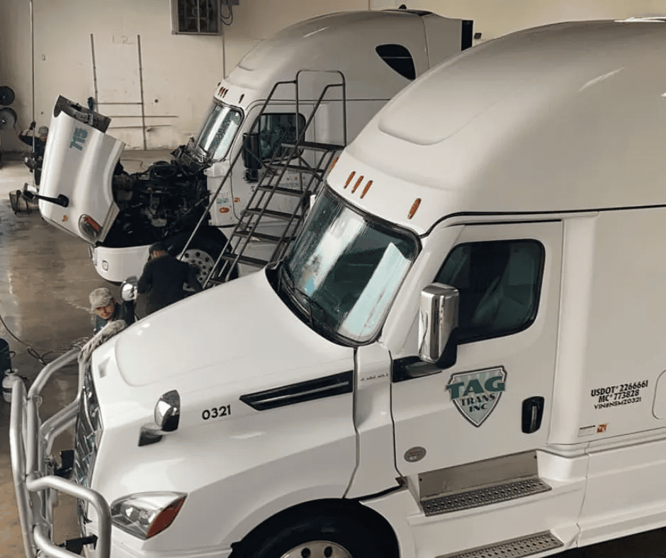 Two white semi-trucks are parked inside a garage. One truck has its hood open with two people working near the engine, while the other truck has its hood closed. Both display the TAG Trans Inc. logo.